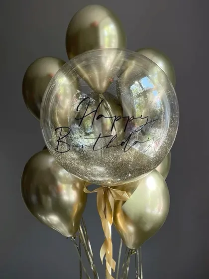 A glittering gold birthday balloon bunch. The arrangement features a personalized clear balloon filled with gold glitter, surrounded by chrome gold helium balloons.