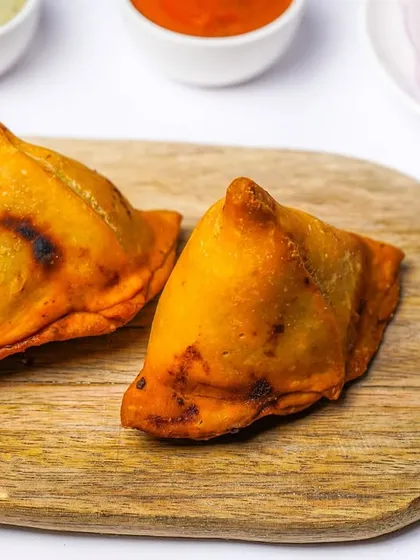 Two baked samosas with a slightly charred, crispy crust, presented simply on a wooden board. This image highlights a healthier alternative to the fried version, shot cleanly for a menu.