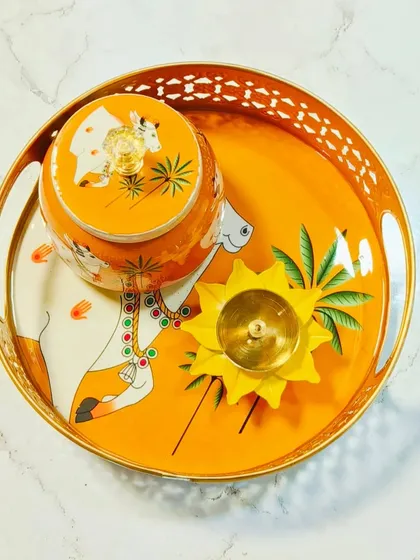 This orange Pichwai tray set features a traditional cow motif, perfect for pooja or as a decorative centerpiece. The set includes a matching jar and a brass diya, blending utility with artistic charm.