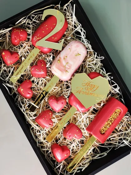 A personalized anniversary gift box designed to celebrate a special milestone. This set features custom-engraved cakesicles, including a number '2' and a 'Happy Anniversary' plaque, paired with heart-shaped chocolates.