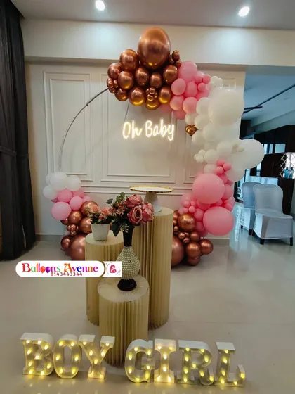 We design beautiful setups for baby showers and gender reveal parties. This arrangement features a circular balloon arch with rose gold, pink, and white balloons, an 'Oh Baby' neon sign, illuminated 'BOY GIRL' marquee letters, and elegant pedestals for cakes or gifts.