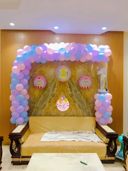 A pastel pink, purple, and blue balloon arch over a sofa for a first birthday party at home.