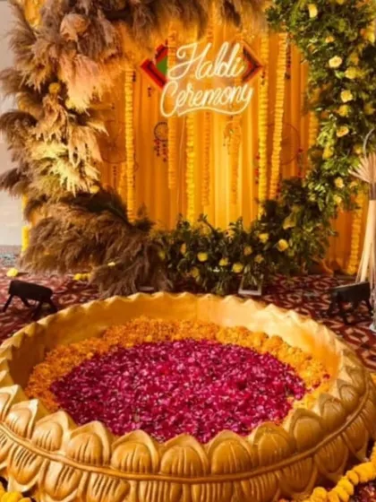A unique Haldi ceremony setup with a large urli filled with red rose petals. The backdrop features a neon sign and is framed by pampas grass and marigold strings for a rustic-chic look.