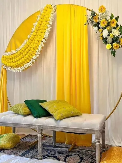A simple and modern Haldi seating area with a circular golden frame. The frame is decorated with yellow drapes and a partial floral arrangement, creating a chic and minimalist look.