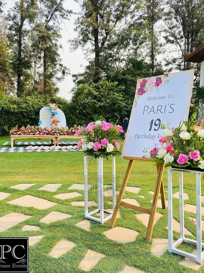 A beautiful outdoor birthday party setup in a lush garden. A personalized welcome sign on an easel, flanked by floral arrangements, greets guests as they arrive for Pari's 19th birthday.