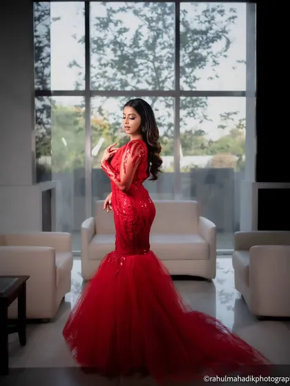 A full-length shot that showcases the dramatic mermaid silhouette and long train of the red gown. The minimalist interior setting keeps the focus entirely on her and the stunning outfit.