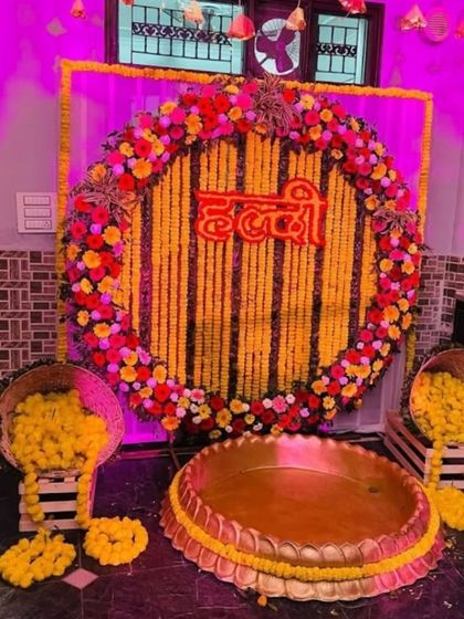 A traditional Haldi backdrop for a home ceremony. The setup features a wall of marigold strings with the word 'Haldi' in Hindi, surrounded by baskets of fresh flowers.