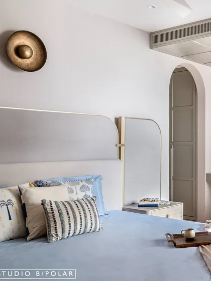 An arched entryway leads to the wardrobe and bathroom, creating a graceful transition. The custom headboard has an asymmetrical design that incorporates a mirror, adding a unique touch.