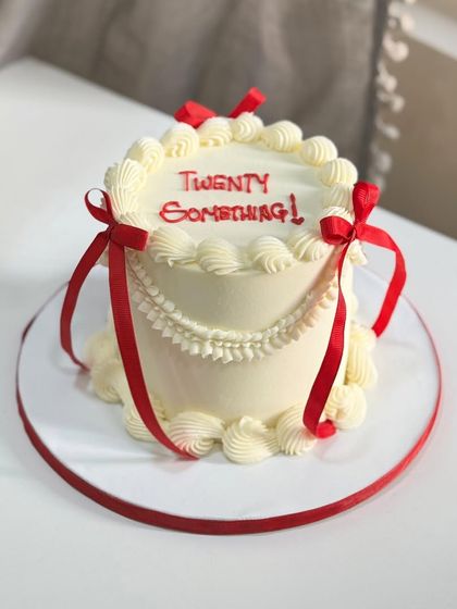 Twenty-something and loving every layer. This classic cake features delicate piping and red ribbons, offering subtle drama and timeless charm for a beautiful phase of life.
