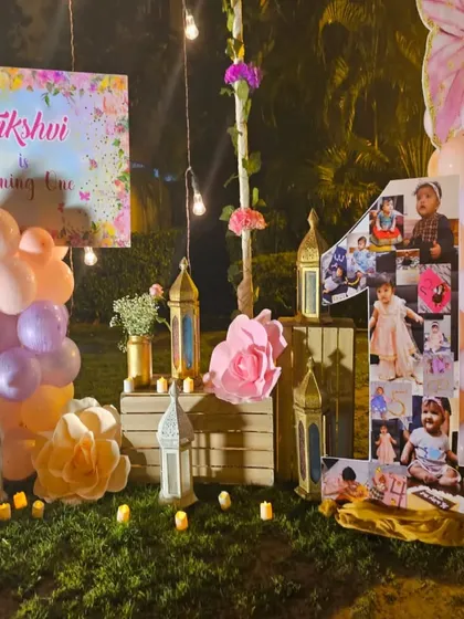 Here is a wider view of an outdoor first birthday party we organized. The setup includes a personalized welcome sign, balloon arrangements, and a beautiful decor corner with a photo collage and lanterns, all lit by charming string lights.