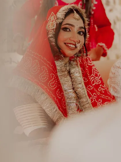 A joyful bride peeking through her red bandhani dupatta, adorned with a traditional nath and maang tikka.