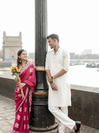 A sweet moment during a pre-wedding shoot at the Gateway of India, where the bride in a red saree holds a bouquet of sunflowers while looking lovingly at her partner.