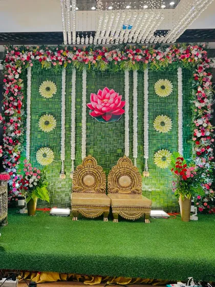 A beautiful stage with a backdrop of woven green leaves and a central lotus motif. The traditional golden chairs and floral arrangements make it perfect for a wedding or naming ceremony.