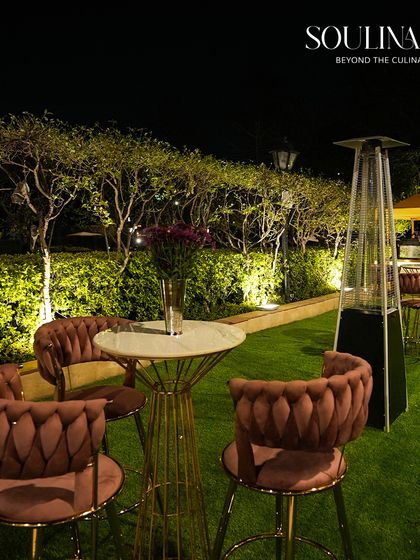 An elegant outdoor soirée setup with high-top tables and plush velvet chairs, set against a backdrop of beautifully lit trees. We design sophisticated spaces for guests to mingle and connect.
