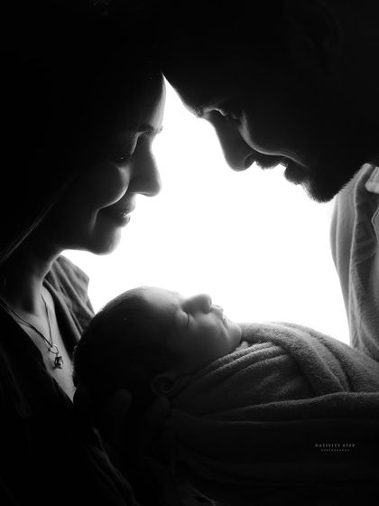 An intimate black and white family portrait. The backlighting creates a beautiful silhouette effect, highlighting the profiles of the parents as they gaze at their newborn.