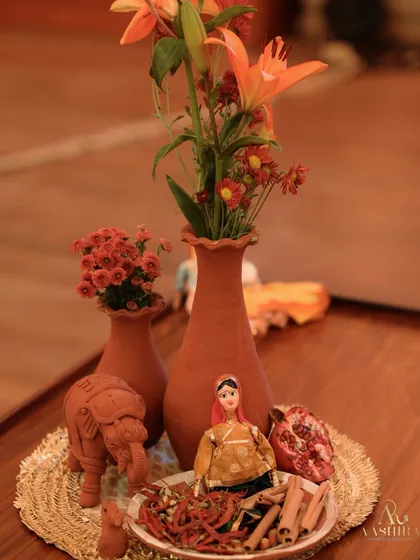 A charming and rustic centerpiece featuring a Rajasthani doll, a clay elephant, and fresh pomegranates, all arranged on a woven mat.