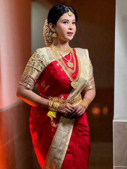 A full portrait of bride Srishti, looking serene and beautiful in her red and cream bridal saree. The traditional jewelry and simple hair bun complete her classic Kerala bridal look.