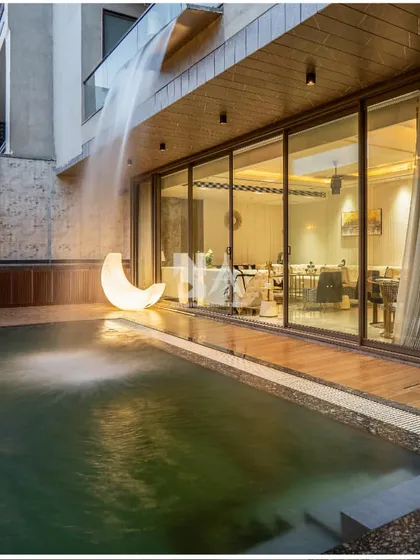 We extend luxury to the outdoors. This poolside area is designed for ultimate relaxation and sophisticated entertaining, featuring a tranquil water curtain cascading from the balcony, a sleek wooden deck, and ambient lighting that transforms the space at dusk.