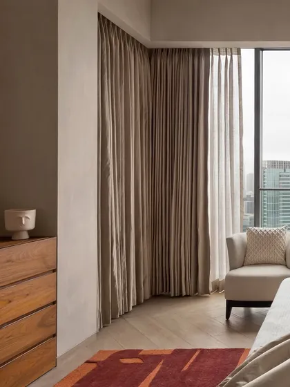 This bedroom in the East Coast Residence is a symphony of warm, earthy tones and tactile materials. The corner window offers expansive city views, while the wooden chest of drawers adds natural warmth to the space.