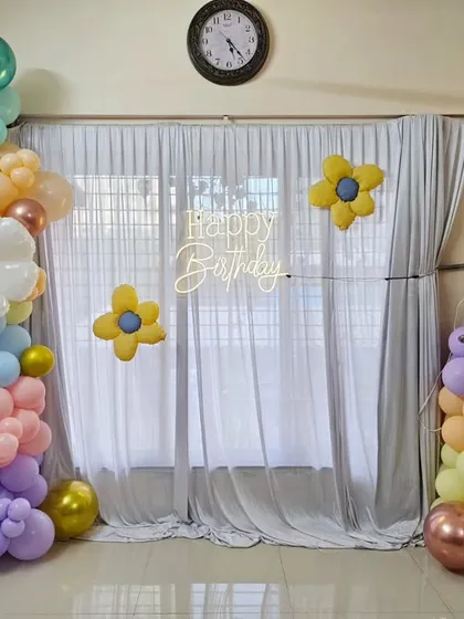 A sunflower-themed first birthday setup with a pastel organic balloon garland, a 'Happy Birthday' neon sign, and a large '1' foil balloon.