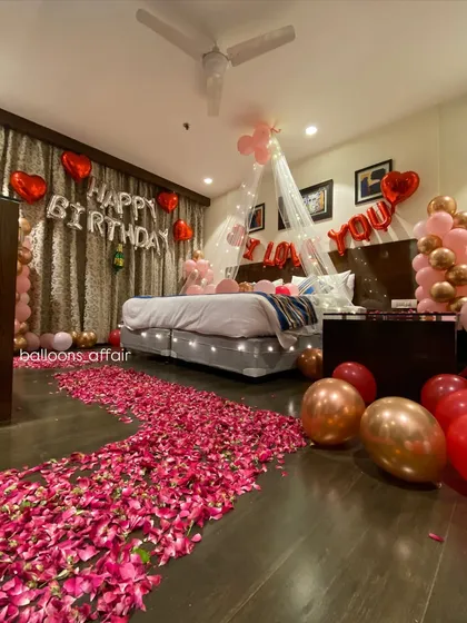 A romantic birthday surprise filling a hotel room with love. The decor includes a rose petal pathway, a canopy with fairy lights over the bed, 'I Love You' and 'Happy Birthday' foil balloons, and balloon arrangements in red and rose gold.