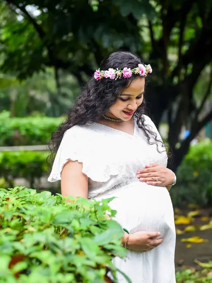 A serene portrait of a mom-to-be in a white dress and flower crown, cradling her bump amidst lush green bushes.