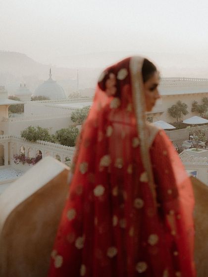 A soft-focus shot of the bride looking out from a palace balcony. The red of her veil against the hazy Udaipur skyline creates a romantic and evocative image.
