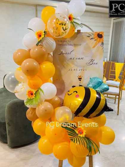 A welcome stand for a bee-themed baby shower. A large bee foil balloon and a cascade of yellow and white balloons create a bright and happy entrance for guests.