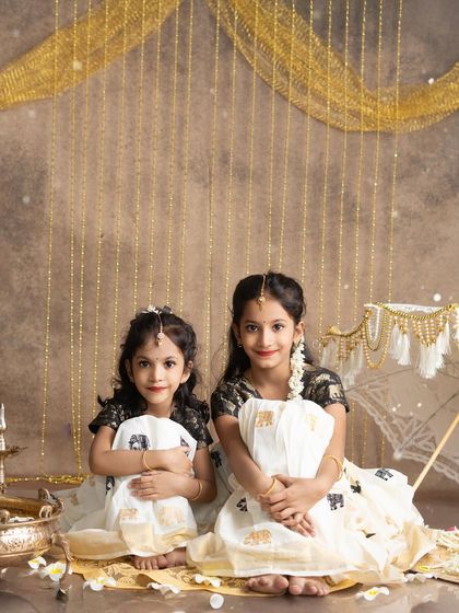A sweet portrait of two sisters sitting together during their Onam photoshoot. The golden backdrop and traditional props create a warm, festive atmosphere.