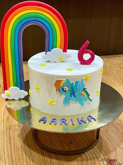 This cake features Rainbow Dash from My Little Pony. The design includes a large fondant rainbow and edible stars, all on a clean white whipped cream base.
