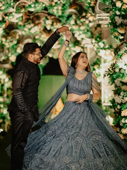 A happy couple enjoying their sangeet night. The bride's twirling grey lehenga and the groom's classic black sherwani are perfectly complemented by the lush green floral backdrop I designed for their photo corner.