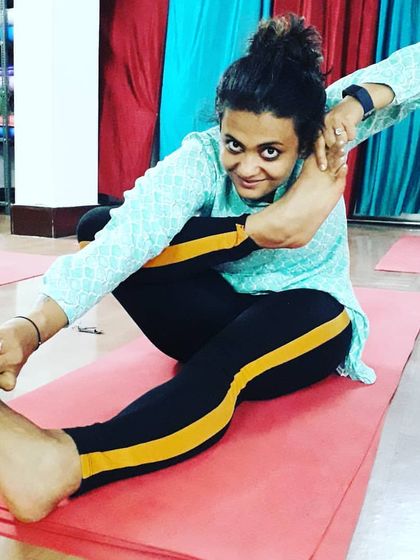 A student smiles while practicing Akarna Dhanurasana (Archer's Pose). This seated posture requires significant hip and hamstring flexibility, demonstrating a joyful approach to challenging asanas.