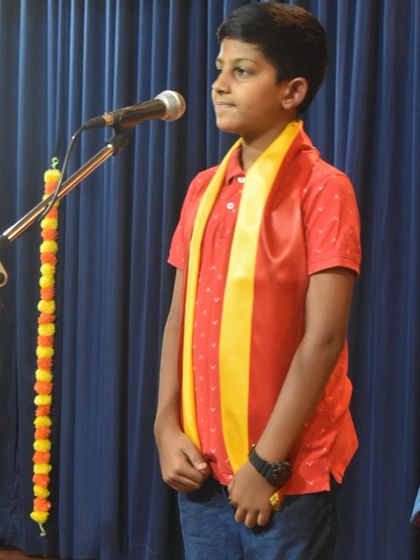 A young student stands confidently at the microphone, ready to perform his piece for the Kannada Rajyotsava celebration.