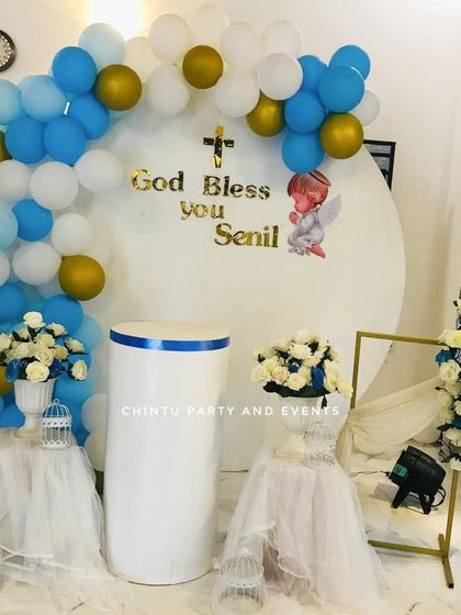 A simple christening decoration for a boy. The setup uses a blue and white balloon arch, tulle-draped tables, and an angel cutout for a blessed event.