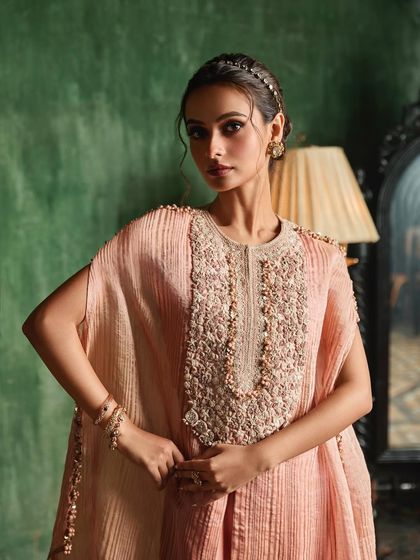 A model poses in a textured pink cape set with a pearl and bead embroidered neckline, exuding regal elegance.