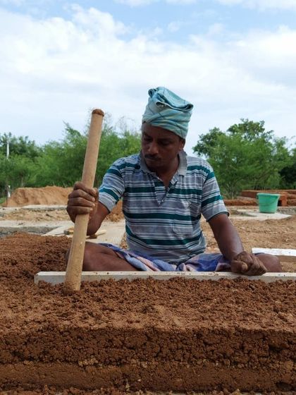 An artisan tamps down a layer of earth while building a cob wall. This ancient technique involves mixing local soil, water, and fibrous material, and then sculpting it by hand to create thick, load-bearing walls that are incredibly strong and thermally efficient.