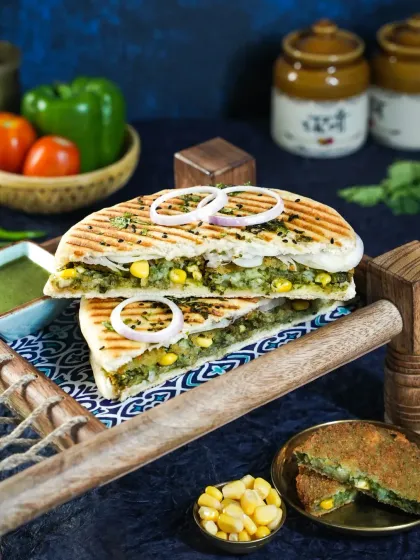 A stacked grilled sandwich filled with corn and spinach, served on a rustic wooden tray. The moody blue background and traditional pickle jars create a fusion of modern cafe food with Indian flavors.