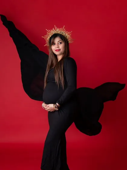 A regal maternity portrait of a woman in a black gown and a golden sunburst crown. The bold red background contrasts with the black dress, creating a powerful and queenly image.
