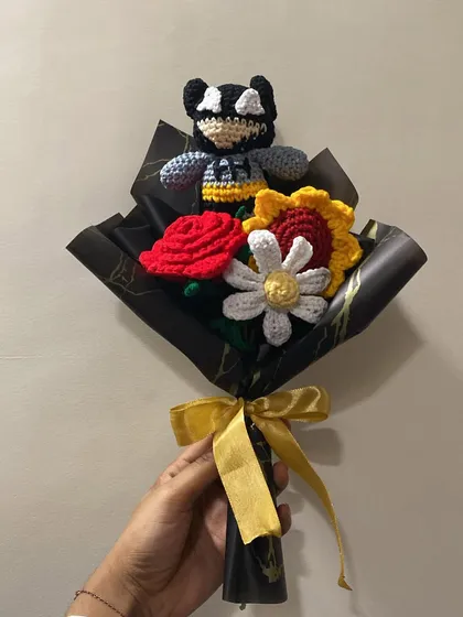 A unique bouquet featuring a Batman-inspired character. The dark wrapping paper with gold accents makes the colors of the crochet rose, sunflower, and daisy pop.