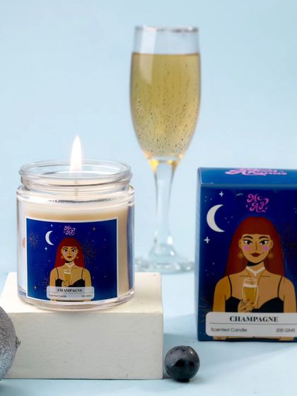 A "Champagne" scented candle is styled with a festive silver ornament and a glass of bubbly. The cool blue background and sparkling elements create a celebratory and sophisticated mood.