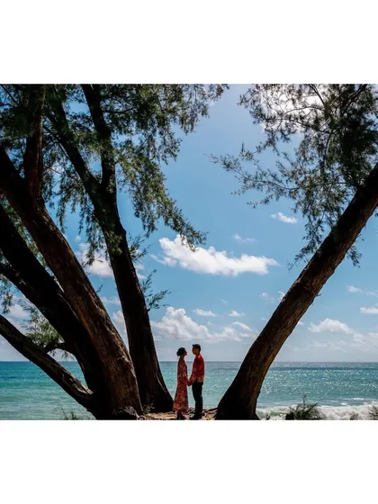 Shachi and Nick stand under the trees on the beach in Phuket. The natural frame created by the trees and the stunning ocean view make this a breathtaking destination wedding portrait.