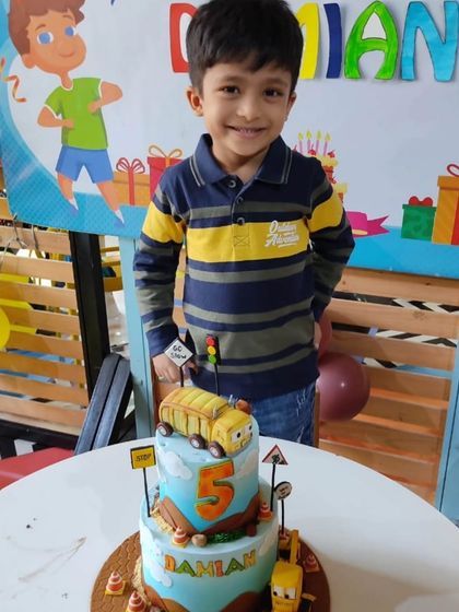 Damian proudly standing by his two-tier "Stinky & Dirty" cake, featuring the show's main characters, a garbage truck and a backhoe loader.