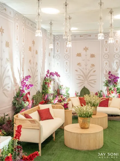 An elegant lounge setup at the entrance, featuring modern cane-back sofas, vibrant red cushions, and lush floral arrangements against a wall with traditional block-print motifs.