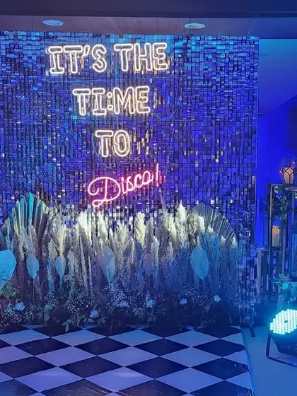 A vibrant and energetic photo booth for a 30th birthday disco party. The setup includes a dazzling sequin wall, a custom neon sign, and fun props, all illuminated by cool blue lighting to get everyone in the mood to dance.