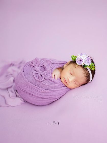 Simply serene. A newborn wrapped in a beautiful purple swaddle against a matching backdrop, showing off the intricate wrap style and a delicate floral headband.