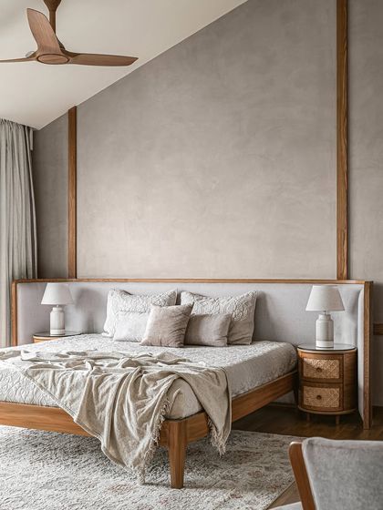 A bedroom with a serene, minimalist design. The space features a low wooden bed with an upholstered headboard, set against a textured grey wall with wood trim. The rattan nightstands add a natural, rustic touch.
