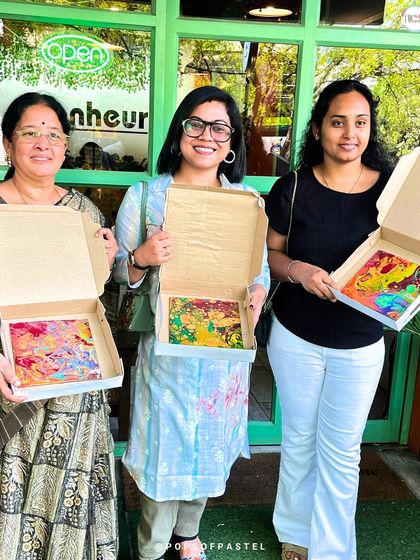 Three women proudly display their finished fluid art pieces from our special Women's Day workshop. It was a day of color, coffee, and connection.