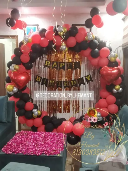 A birthday setup in a living room, featuring a circular backdrop made of red and black balloons. The center table is covered with rose petals, adding a touch of romance and color to the decor.