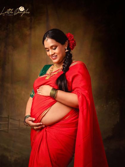 A serene moment of a mom-to-be cradling her bump. The rich red saree and the fine art backdrop create a warm and traditional feel.
