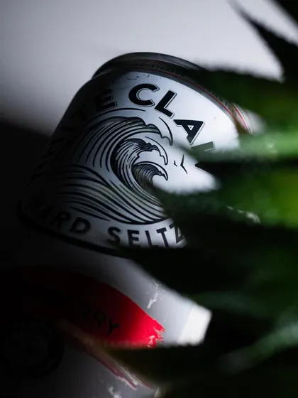 An artistic, creative shot of a White Claw can, partially obscured by a plant. This style of product photography is intriguing and modern.